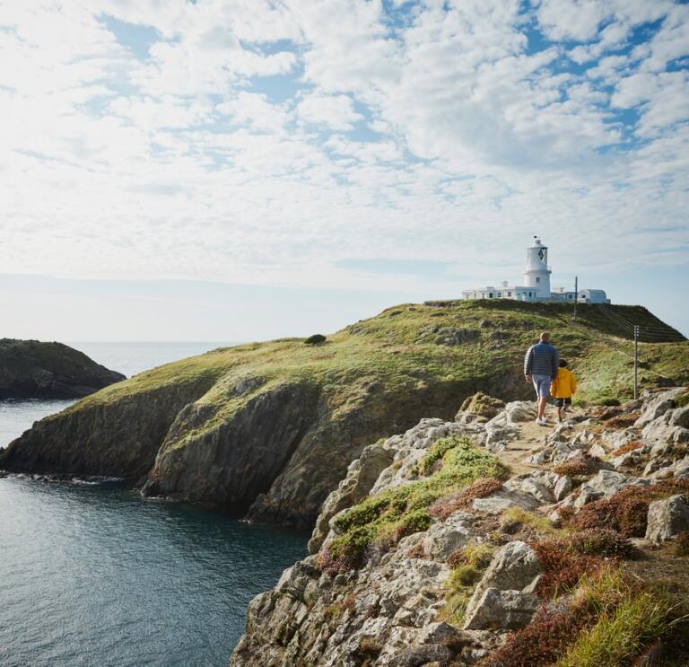 Two people walking along the cliffs towards a lighthouse.